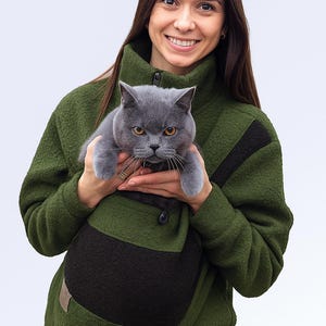 May include: A grey cat is held in a green fleece jacket with a black pocket. The jacket has a high collar and a button closure. The cat has yellow eyes and is looking at the camera. The background is white.