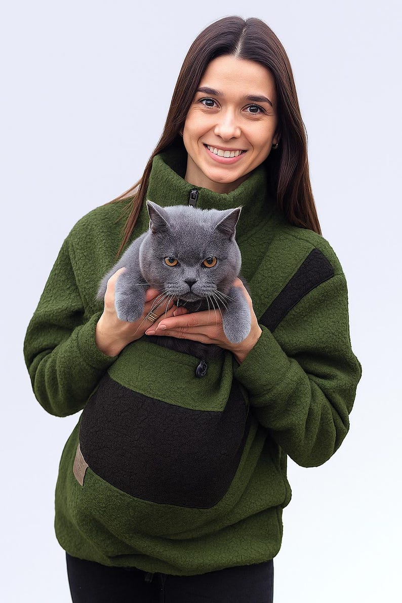 May include: A grey cat is held in a green fleece jacket with a black pocket. The jacket has a high collar and a button closure. The cat has yellow eyes and is looking at the camera. The background is white.
