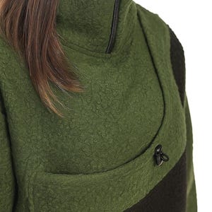 May include: Green fleece jacket with a high collar and a black zip. The jacket has a black stripe across the chest and a small black toggle detail. The fabric has a textured, nubby appearance.