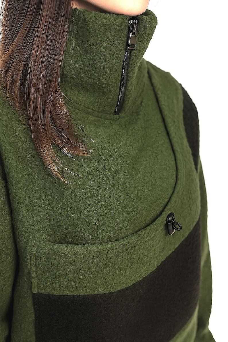 May include: Green fleece jacket with a high collar and a black zip. The jacket has a black stripe across the chest and a small black toggle detail. The fabric has a textured, nubby appearance.