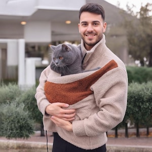 Peut inclure: Veste polaire beige avec un porte-chat intégré marron et beige. Un chat gris aux yeux orange est niché dans le porte-chat. La veste a un col montant et une fermeture éclair.