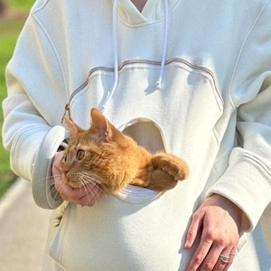 May include: A white hoodie with a large pocket on the front that is designed to hold a cat. A ginger cat is sitting in the pocket.