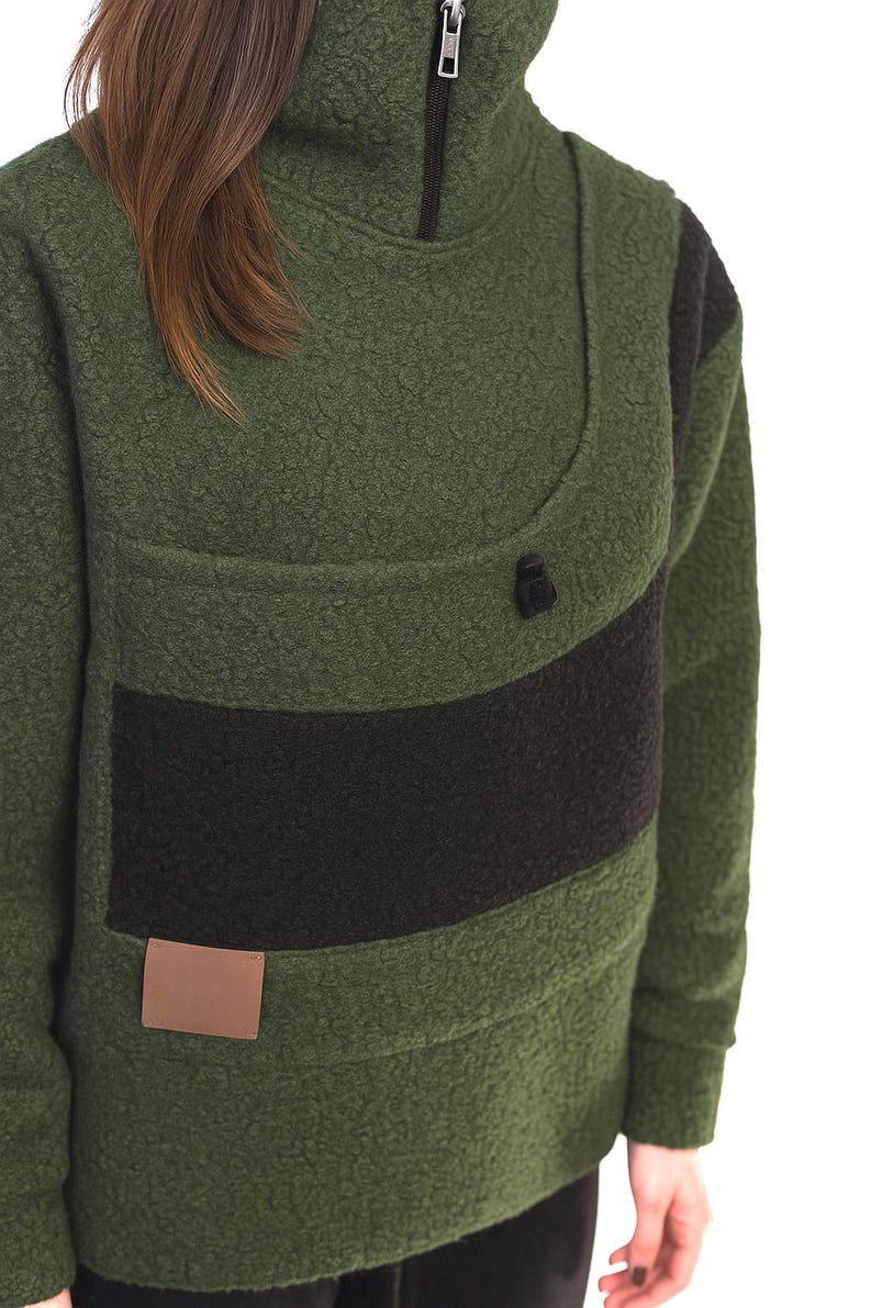 May include: Green fleece pullover with a high collar and a black zip. The pullover has a black horizontal stripe across the chest and a small brown rectangular patch near the bottom. The fabric has a textured, nubby appearance.