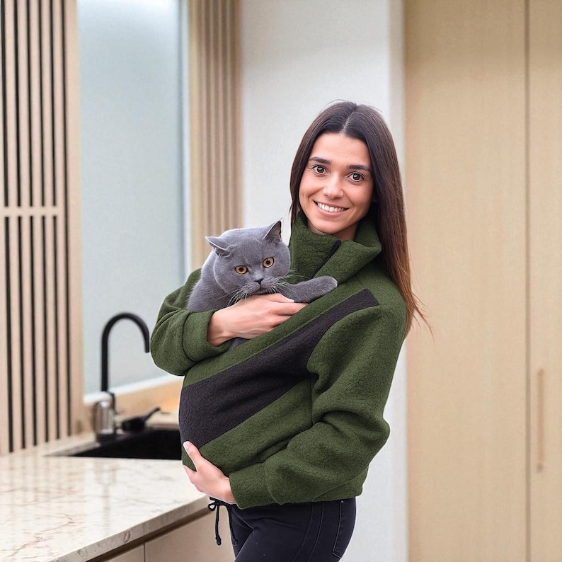 May include: A green and black pet carrier jacket holds a grey cat. The jacket has a high collar and a front pocket designed to carry a cat. The person holding the jacket is smiling.