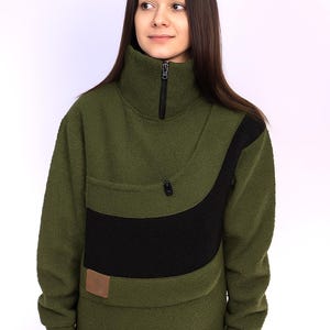 May include: Olive green fleece pullover with a high collar and a black accent panel. The pullover features a zippered neckline, a small pocket, and a brown rectangular patch. The garment is designed for warmth and comfort.