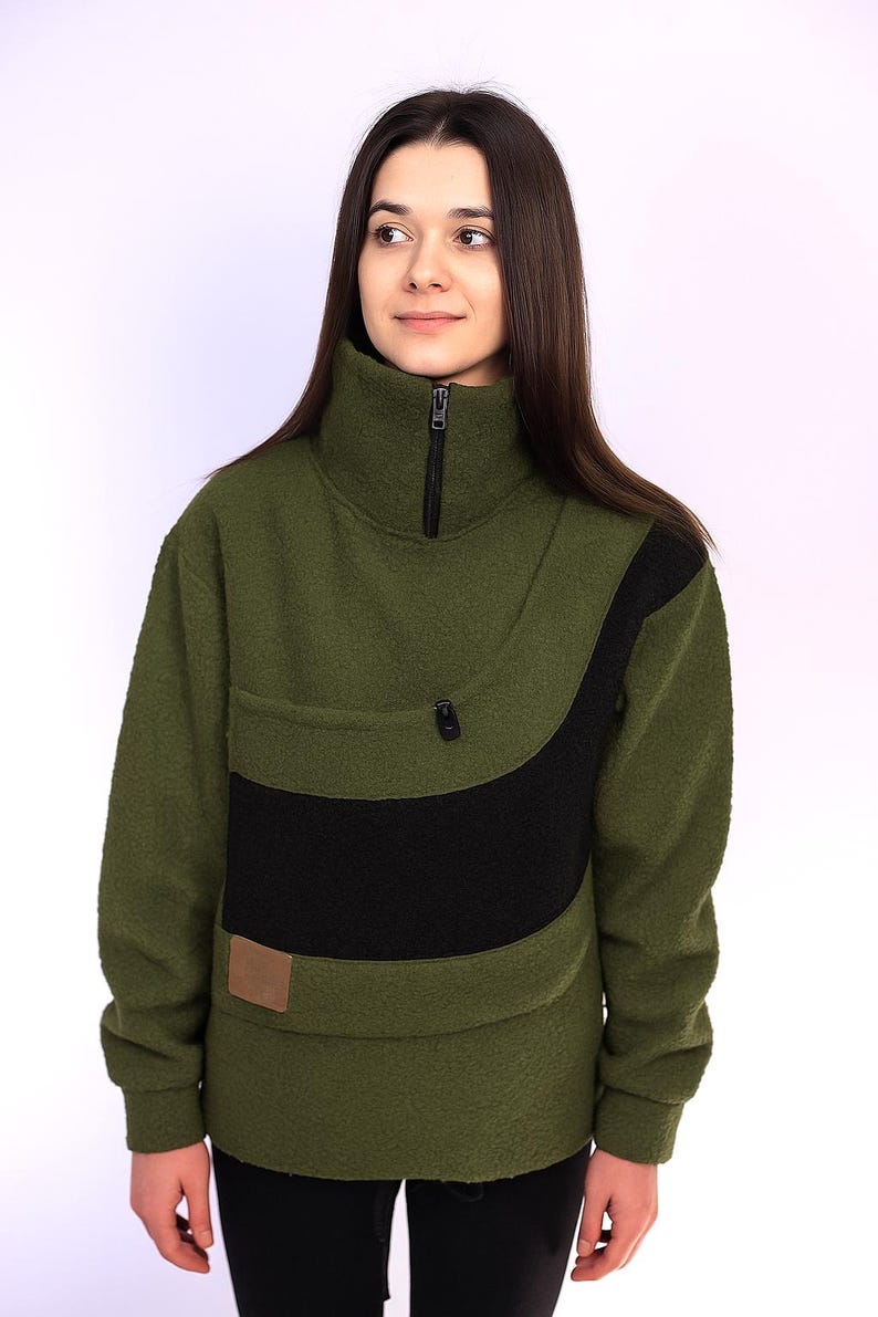 May include: Olive green fleece pullover with a high collar and a black accent panel. The pullover features a zippered neckline, a small pocket, and a brown rectangular patch. The garment is designed for warmth and comfort.