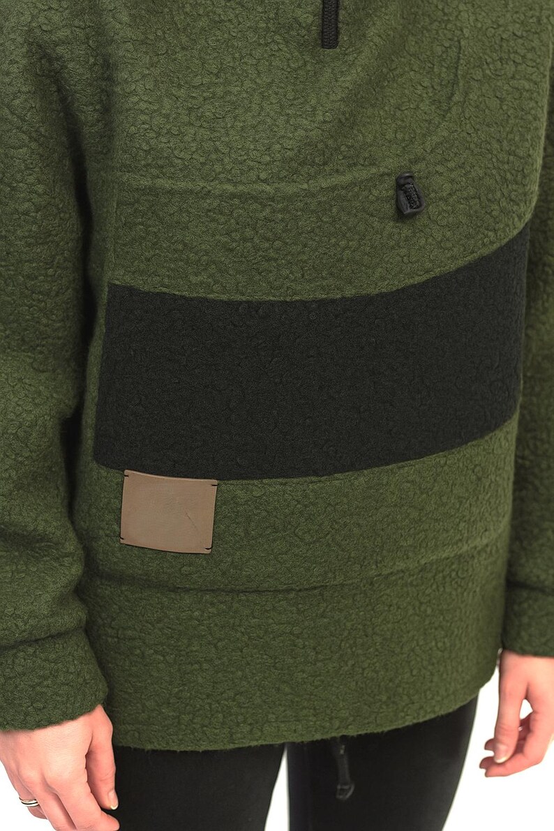 May include: Green fleece pullover with a black stripe across the chest. The pullover features a half-zip and a small, square patch on the lower front. The fabric has a textured, fuzzy appearance. Part of a clothing collection.