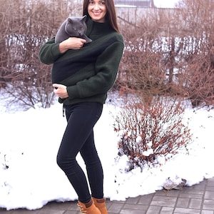 May include: A dark green cat carrier sweater with a black interior. The sweater is worn with black trousers and tan boots. A grey cat is held inside. The background shows snow and winter foliage. The person is smiling.