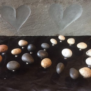 May include: A collection of smooth, round, natural stone drawer pulls in various shades of gray, brown, and white. The pulls are arranged on a black surface.