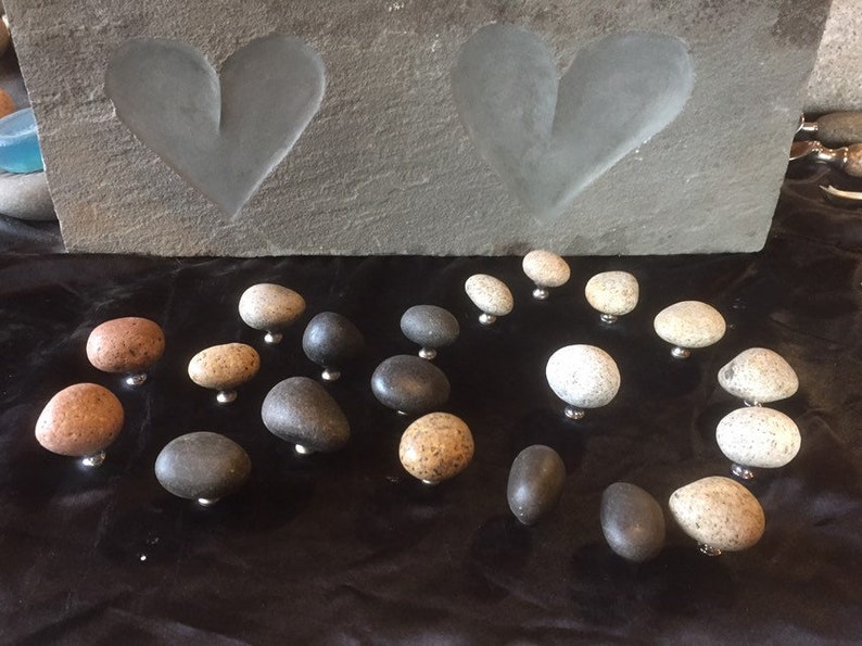 May include: A collection of smooth, round, natural stone drawer pulls in various shades of gray, brown, and white. The pulls are arranged on a black surface.