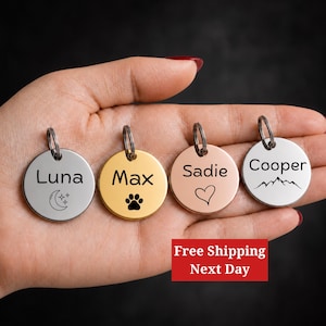 May include: Four circular metal pet ID tags with split rings. The first is silver with the name Luna and a moon icon. The second is gold with the name Max and a paw print. The third is rose gold with the name Sadie and a heart. The fourth is silver with the name Cooper and a mountain graphic.