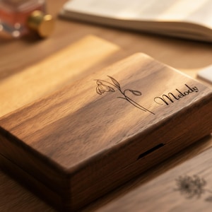 May include: A rectangular wooden box with a floral design and the name "Melody" engraved on the lid. The box is made of dark wood and has a natural wood grain. The floral design is a delicate line drawing of a flower.