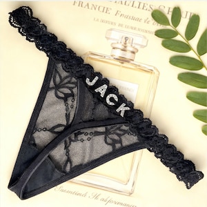 May include: A black lace thong featuring a scalloped waistband adorned with silver-toned rhinestone letters spelling out the word JACK. The garment is crafted from sheer floral patterned lace with solid black trim along the edges and seams.