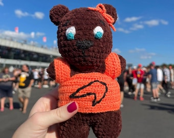May include: A handmade brown crochet bear with an orange shirt featuring a black graphic. The bear has blue and white eyes, a black nose, and an orange bow. The bear is being held in front of a crowd of people.