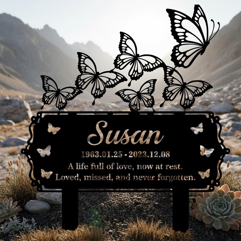 Pu&ograve; includere: Paletto commemorativo da giardino in metallo nero con il nome "Susan", date e un'iscrizione. Il design include ritagli di farfalle. Lo sfondo mostra una catena montuosa.
