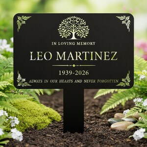 Puede incluir: Una estaca de jard&iacute;n conmemorativa negra con el nombre "LEO MARTINEZ" y las fechas 1939-2026 en letras doradas. La estaca tiene un dise&ntilde;o de &aacute;rbol y las palabras "IN LOVING MEMORY" y "ALWAYS IN OUR HEARTS AND NEVER FORGOTTEN".