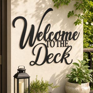 Könnte beinhalten: Ein schwarzes Metall-Wandschild mit dem kursiven Schriftzug Welcome to the Deck. Das Schild ist an einer hellen Wand über einer schwarzen Laterne und einer Topfpflanze angebracht. Die Schrift ist fett und dunkel und bildet einen Kontrast.