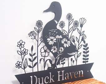 Puede incluir: Silueta de metal negro de un pato con flores y el texto "Duck Haven" en una pancarta negra.