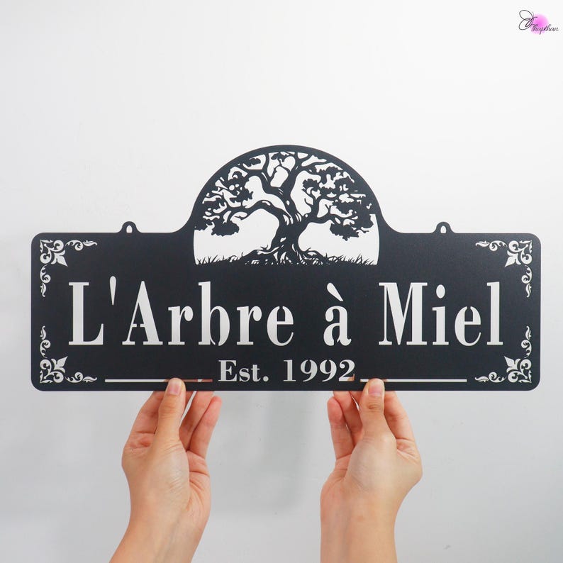 May include: Black metal sign with the text "L'Arbre &agrave; Miel" and "Est. 1992" in white. The sign features a tree silhouette at the top. The sign is held by two hands against a white background.