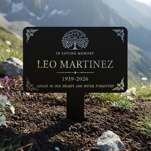 Puede incluir: Una estaca de jard&iacute;n conmemorativa negra con el texto "IN LOVING MEMORY LEO MARTINEZ 1939-2026 ALWAYS IN OUR HEARTS AND NEVER FORGOTTEN." La estaca presenta un dise&ntilde;o de &aacute;rbol y detalles florales. Se coloca en el suelo con un fondo monta&ntilde;oso.