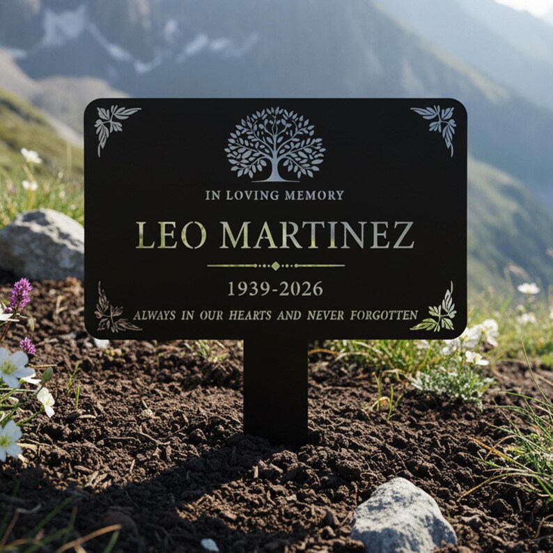 Puede incluir: Una estaca de jard&iacute;n conmemorativa negra con el texto "IN LOVING MEMORY LEO MARTINEZ 1939-2026 ALWAYS IN OUR HEARTS AND NEVER FORGOTTEN." La estaca presenta un dise&ntilde;o de &aacute;rbol y detalles florales. Se coloca en el suelo con un fondo monta&ntilde;oso.