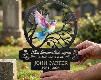 Puede incluir: Estaca conmemorativa de metal negro con un colibrí colorido dentro de un marco circular. La estaca incluye el texto "When hummingbirds appear a love one is near" y el nombre "JOHN CARTER 1964-2025".