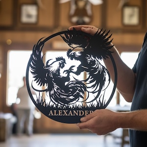 Puede incluir: Arte de pared de metal negro con dos gallos luchando en un círculo. El nombre "ALEXANDER" está en la parte inferior. Esta pieza decorativa es adecuada para uso en interiores o exteriores. El diseño también muestra hierba.
