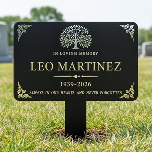 Puede incluir: Un marcador conmemorativo negro con letras doradas y un dise&ntilde;o de &aacute;rbol. El texto dice: "IN LOVING MEMORY LEO MARTINEZ 1939-2026 ALWAYS IN OUR HEARTS AND NEVER FORGOTTEN." El marcador est&aacute; en hierba verde.