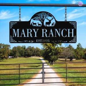 May include: Black metal sign with the text "MARY RANCH" and "EST.2025". The sign features a silhouette of deer and a tree. The sign is hanging from a metal bar with chains. The background shows a ranch entrance.