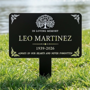 Puede incluir: Estaca de jard&iacute;n conmemorativa negra con el texto "IN LOVING MEMORY LEO MARTINEZ 1939-2026 ALWAYS IN OUR HEARTS AND NEVER FORGOTTEN." Presenta un gr&aacute;fico de &aacute;rbol y detalles decorativos en las esquinas. Colocada en hierba verde cerca del agua.
