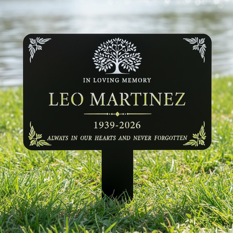 Puede incluir: Estaca de jard&iacute;n conmemorativa negra con el texto "IN LOVING MEMORY LEO MARTINEZ 1939-2026 ALWAYS IN OUR HEARTS AND NEVER FORGOTTEN." Presenta un gr&aacute;fico de &aacute;rbol y detalles decorativos en las esquinas. Colocada en hierba verde cerca del agua.