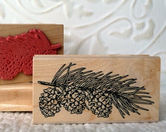 May include: Two wooden stamps with botanical designs. One stamp features a red pine branch impression, while the other displays a black and white illustration of pine cones and needles. The stamps are on a white surface.