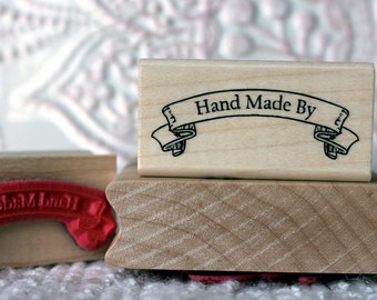 May include: Two wooden stamps with the text "Hand Made By" on a banner. The top stamp has a white banner with black text. The bottom stamp has a red banner with white text.