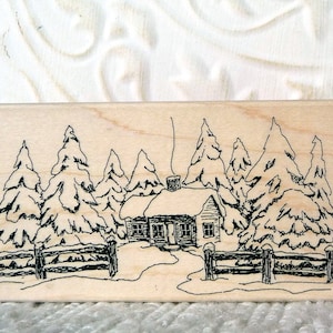 May include: Two wooden mounted rubber stamps with a winter scene of a cabin in the woods. The first stamp is a black and white line drawing of a cabin with snow-covered trees and a fence. The second stamp is a red rubber stamp of the same scene.
