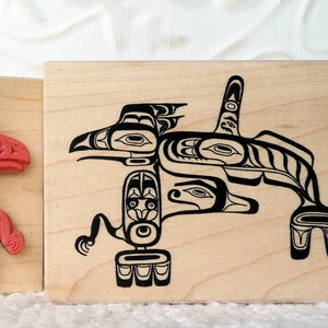 May include: Two wooden blocks with carved designs of Native American art. The left block has a red design of a bird with a fish in its beak. The right block has a black design of a bird with a fish in its beak.