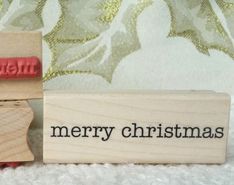 May include: Three wooden rubber stamps with red ink. The top stamp reads "Christmas Greetings". The middle stamp is blank. The bottom stamp reads "merry christmas".