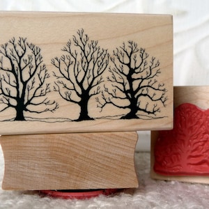 May include: Three wooden mounted rubber stamps with tree designs. The first stamp features three bare trees with black silhouettes on a natural wood background. The second stamp features three red detailed trees on a natural wood background.