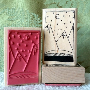 May include: Two wooden stamps with a mountain and star design. The top stamp is a light brown wood with a black and white design of mountains and stars. The bottom stamp is a darker brown wood with a red design of mountains and stars.