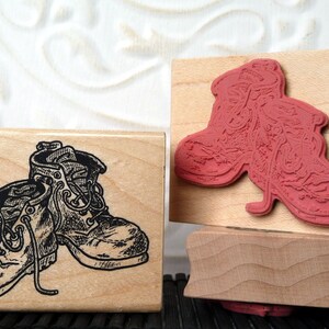 May include: Two wooden rubber stamps. One stamp features a detailed black and white illustration of a pair of hiking boots. The other stamp has a red rubber boot design. The stamps are on a white background.