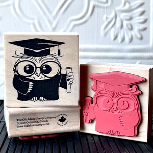 May include: Two wooden rubber stamps featuring a cartoon owl wearing a graduation cap and holding a diploma. The owl on the top stamp is black and white, while the owl on the bottom stamp is pink.