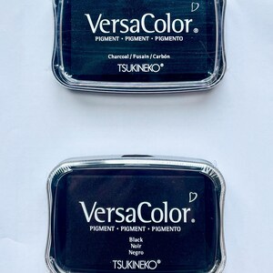 May include: Two black ink pads with clear plastic cases. The top pad says "VersaColor. Pigment Pigment Pigmento Charcoal/Fusain/Carb&oacute;n TSUKINEKO&reg;". The bottom pad says "VersaColor. Pigment Pigment Pigmento Black Noir Negro TSUKINEKO&reg;".