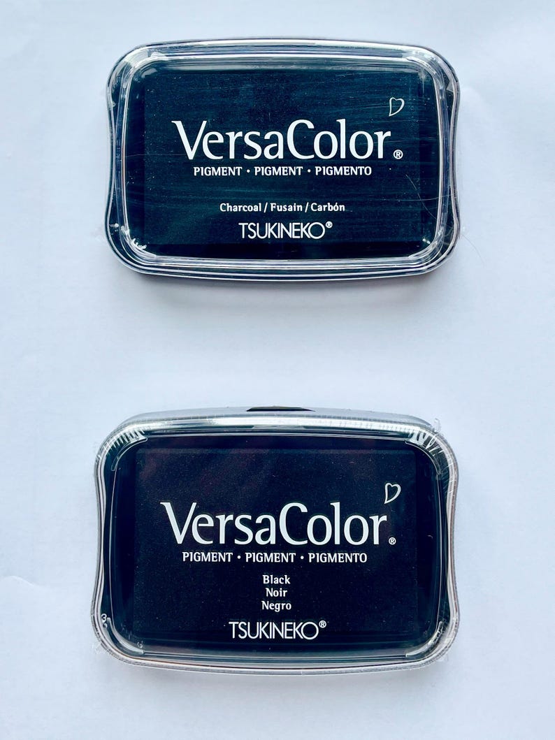 May include: Two black ink pads with clear plastic cases. The top pad says "VersaColor. Pigment Pigment Pigmento Charcoal/Fusain/Carb&oacute;n TSUKINEKO&reg;". The bottom pad says "VersaColor. Pigment Pigment Pigmento Black Noir Negro TSUKINEKO&reg;".