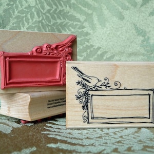 May include: Two wooden rubber stamps with blank rectangular frames. The top stamp has a decorative border with a red background. The bottom stamp has a black and white illustration of a bird and a leafy branch.