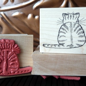 Puede incluir: Dos bloques de madera con sellos de goma. Un sello presenta un gato rojo con un diseño floral. El otro sello presenta un dibujo en blanco y negro de un gato sentado con la cola enroscada alrededor de su cuerpo.