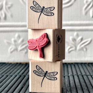 May include: Three wooden blocks featuring a dragonfly design. The top and bottom blocks depict a black dragonfly with intricate details. The middle block showcases a red dragonfly with intricate details. The blocks are stacked on top of one another.
