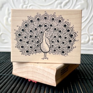 Puede incluir: Un sello de madera con una ilustración en blanco y negro de un pavo real. Las plumas de la cola del pavo real están desplegadas. El sello rectangular está encima de otro sello de madera.
