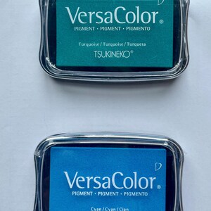 May include: Two ink pads, one turquoise and one cyan, with the text "VersaColor" and "PIGMENT PIGMENT PIGMENTO" and "TSUKINEKO&reg;" on the front.