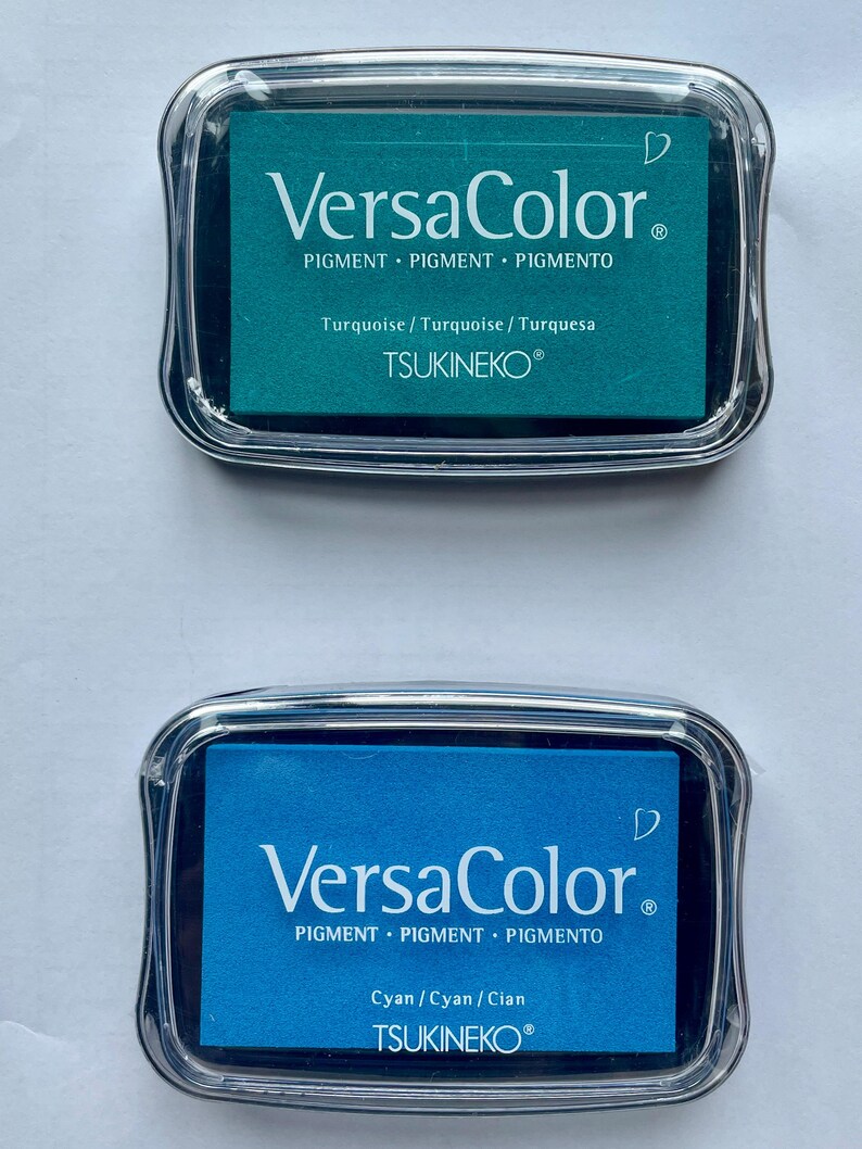 May include: Two ink pads, one turquoise and one cyan, with the text "VersaColor" and "PIGMENT PIGMENT PIGMENTO" and "TSUKINEKO&reg;" on the front.
