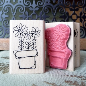 May include: Two wooden rubber stamps with floral designs. The stamp on the left has a black and white design of three flowers in a pot. The stamp on the right has a pink design of three flowers in a pot. Both stamps have the text "AVA-333" on the side.