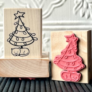 May include: Two wooden stamps featuring a cat sleeping under a Christmas tree with lights. The stamp on the left is black and white and the stamp on the right is red.